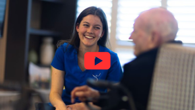 lA woman in a blue uniform smiles while talking to an older man in a chair. A play button icon is overlaid on the image.